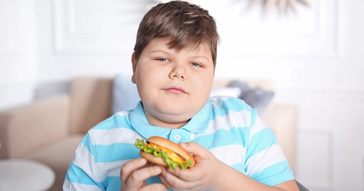 niño con sobrepeso con una hamburguesa ultra procesada en la mano