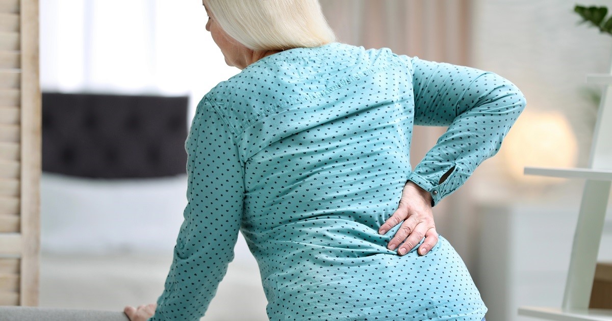 mujer mayor con la mano izquierda se apoya en una silla y con la mano de la derecha se toca las lumbares. Se la ve con signos de presentar dolor lumbar crónico.
