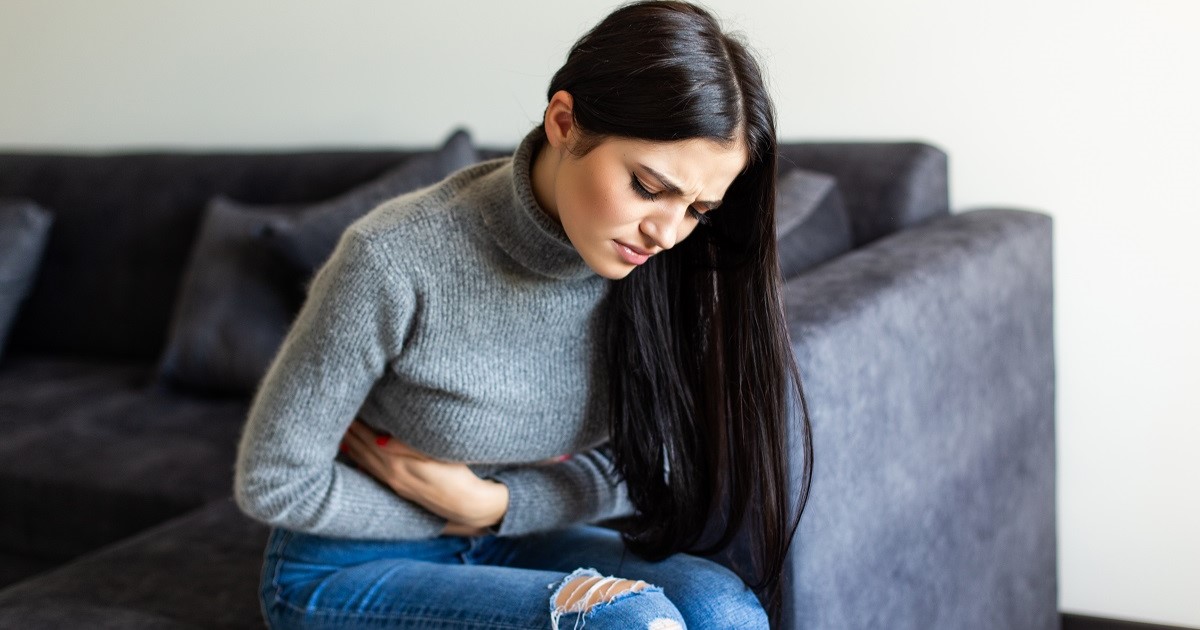 mujer sentada en el sofa encogida, tocándose la barriga con malestar, con posible enfermedad digestiva