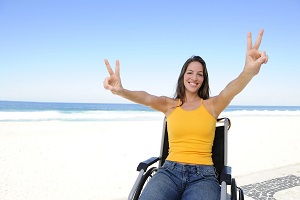 Una joven en silla de ruedas en la playa con las manos mostrando el signo de la victoria y disfrutando de las emociones de humor y felicidad