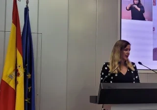 Ana Dávila, consejera de Familia, Juventud y Asuntos Sociales de la Comunidad de Madrid, subida a un atril dando un discurso con las banderas de la Comunidad de Madrid, de España y de la Unión Europea de fondo.